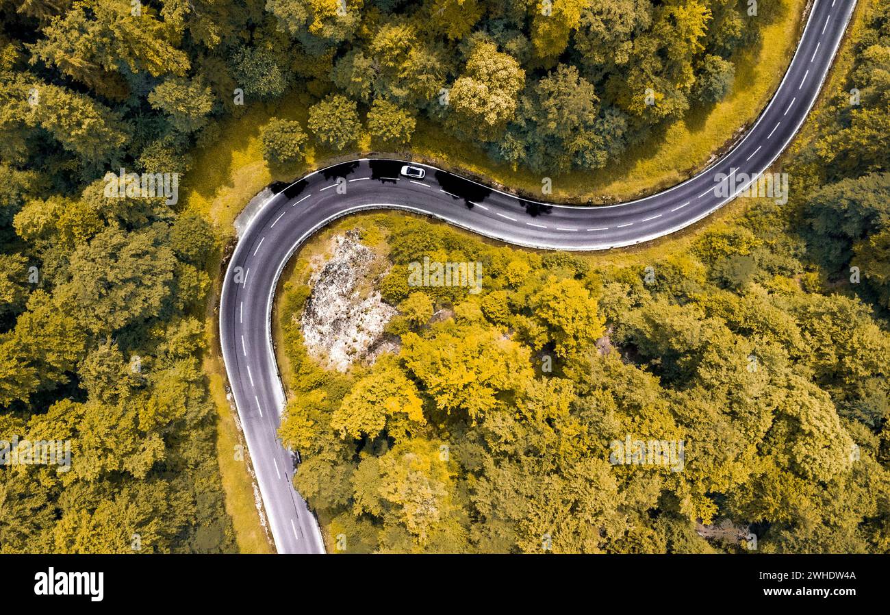 Luftaufnahme einer kurvenreichen Passstraße im Naturpark Altmühltal in Oberbayern. Die Straße schlängelt sich malerisch durch einen sommerlich gemischten Laubwald an einem Felsvorsprung vorbei. Ein weißes Auto fährt um die Kurve. Stockfoto