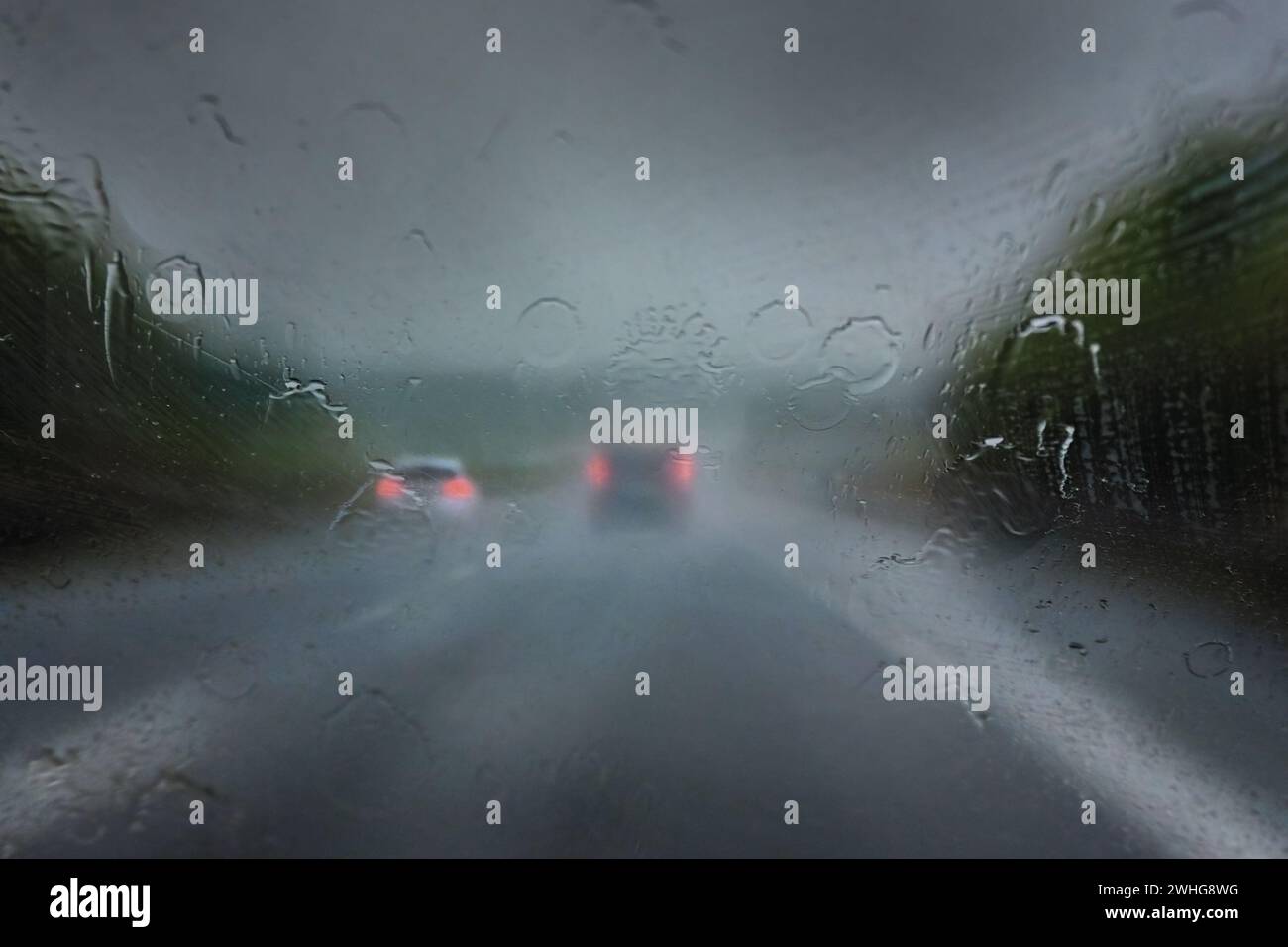 Starker Regen auf der Autobahn, dicke Tropfen auf der Windschutzscheibe und verschwommene Autos mit roten Rücklichtern auf der dunklen Straße, gefährliches Wetter Stockfoto