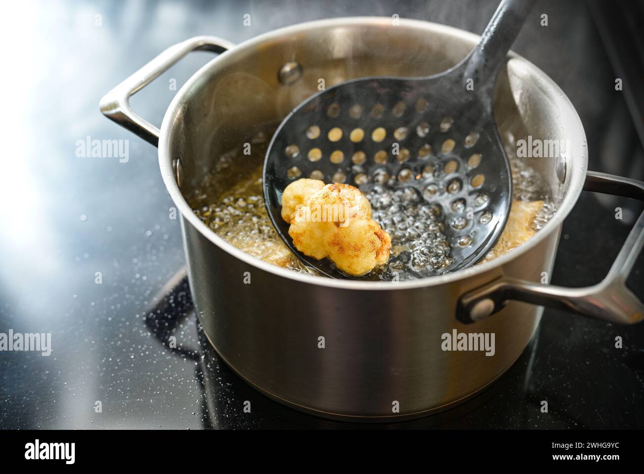 Blumenkohlblume auf einem Spachtel in einem Topf frittiert mit dampfendem Speiseöl auf dem Herd, kreative Zubereitung eines Gemüses Stockfoto
