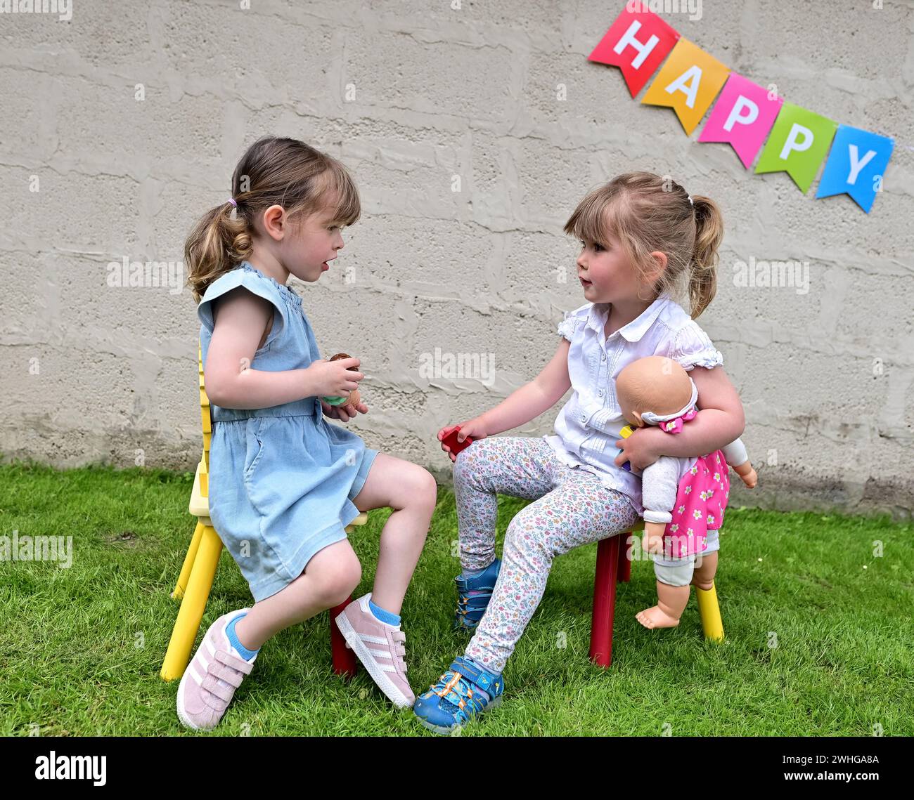 Zwei 3-jährige Kinder sprechen (Mädchen), während sie auf Stühlen auf Gras in einem Garten sitzen, von Angesicht zu Angesicht mit dem Wort HAPPY auf einer weißen Wand dahinter. Stockfoto