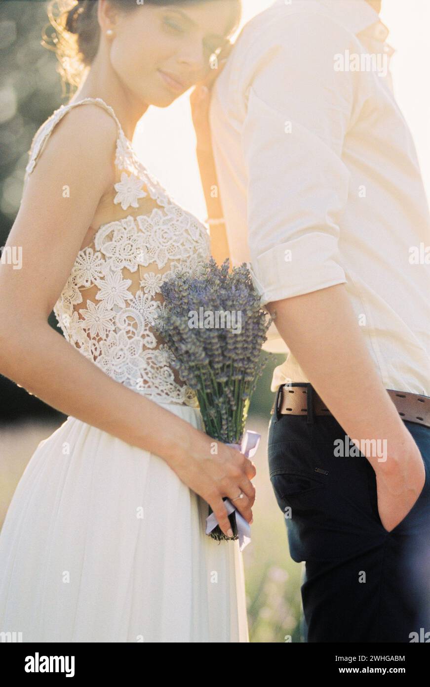 Braut mit einem Lavendelstrauß, der sich gegen den Rücken des auf dem Feld stehenden Bräutigams lehnt Stockfoto