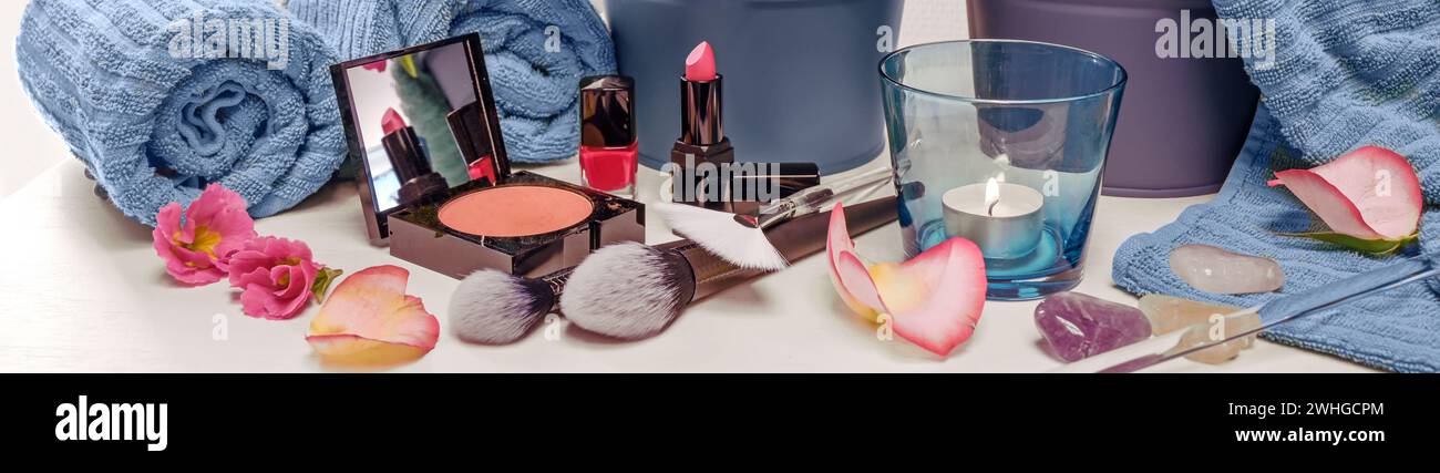 Schminkutensilien wie Gesichtspuder, rosa Lippenstift und nagellack mit Kosmetikbürsten, blauen Handtüchern, Kerze und Blütenblatt Stockfoto