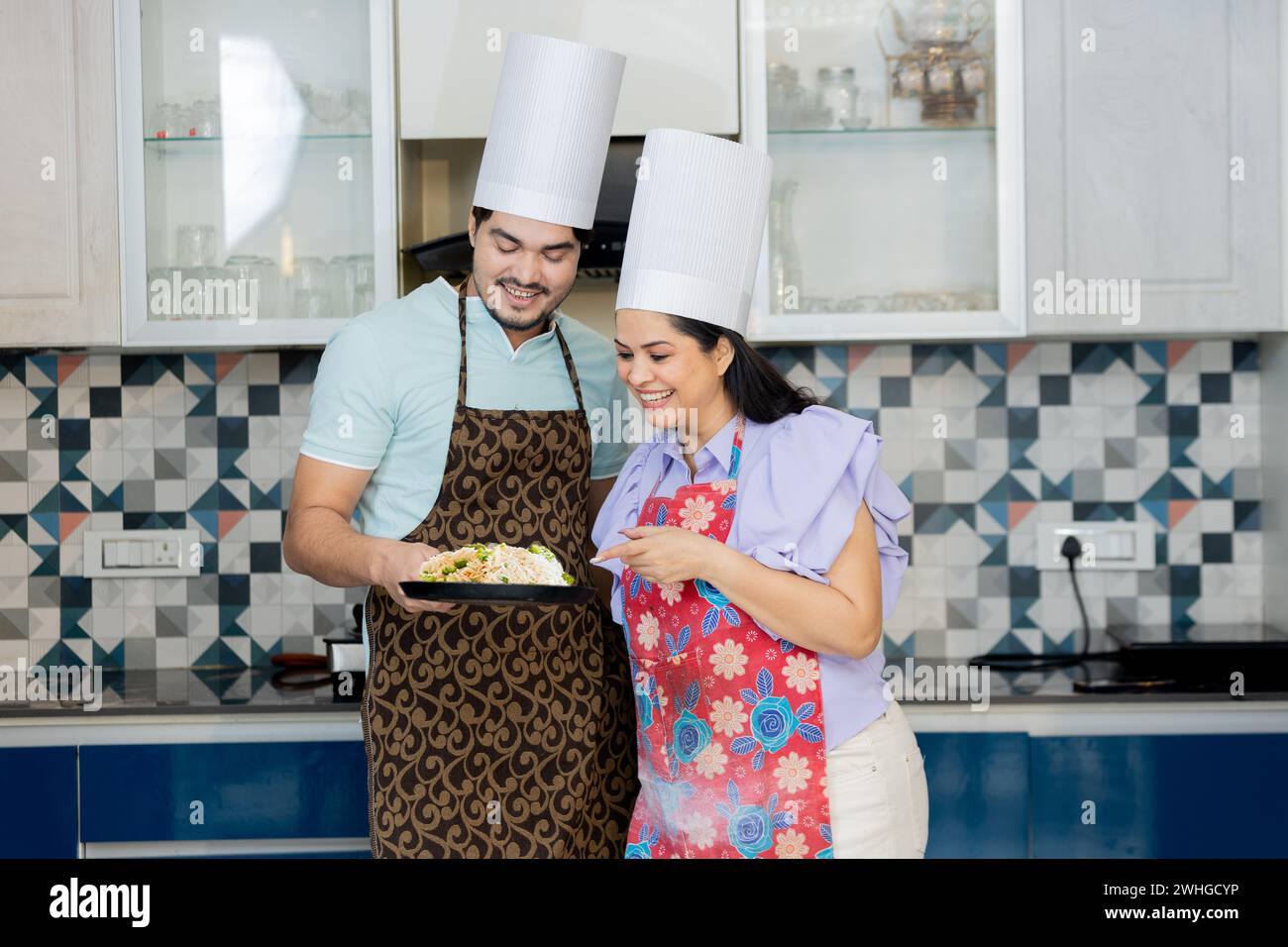 Glückliches indisches Paar in Kochuniform mit Schürze, das leckeres Essen in der Küche zubereitete. Kochfähigkeiten Zeigen Stockfoto