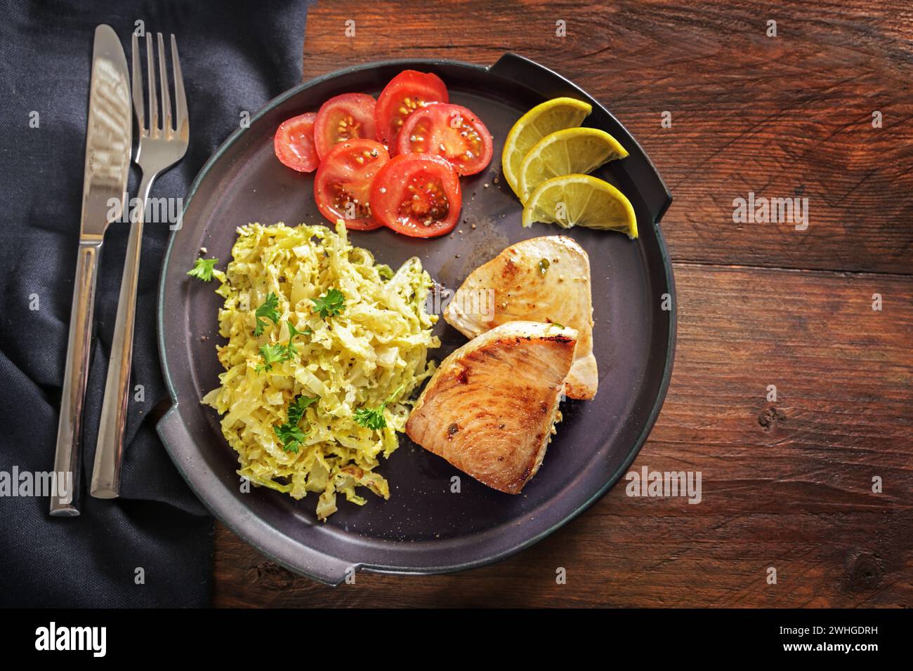 Gebratene Thunfischsteaks mit wirsingkohl, Zitrone und Tomaten auf einem dunklen Teller und ein rustikaler Holztisch mit Serviette und Besteck, Kopie Stockfoto