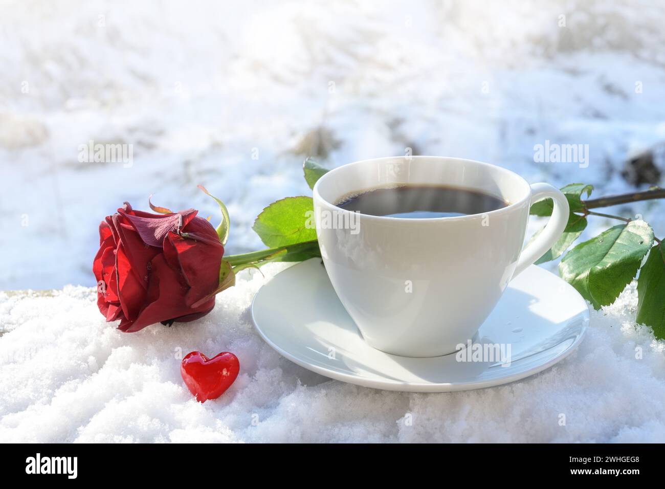 Heißer Kaffee in einer weißen Tasse serviert draußen im Schnee, dekoriert mit einer roten Rose und einem kleinen Herz aus Glas an einem sonnigen Winter Stockfoto