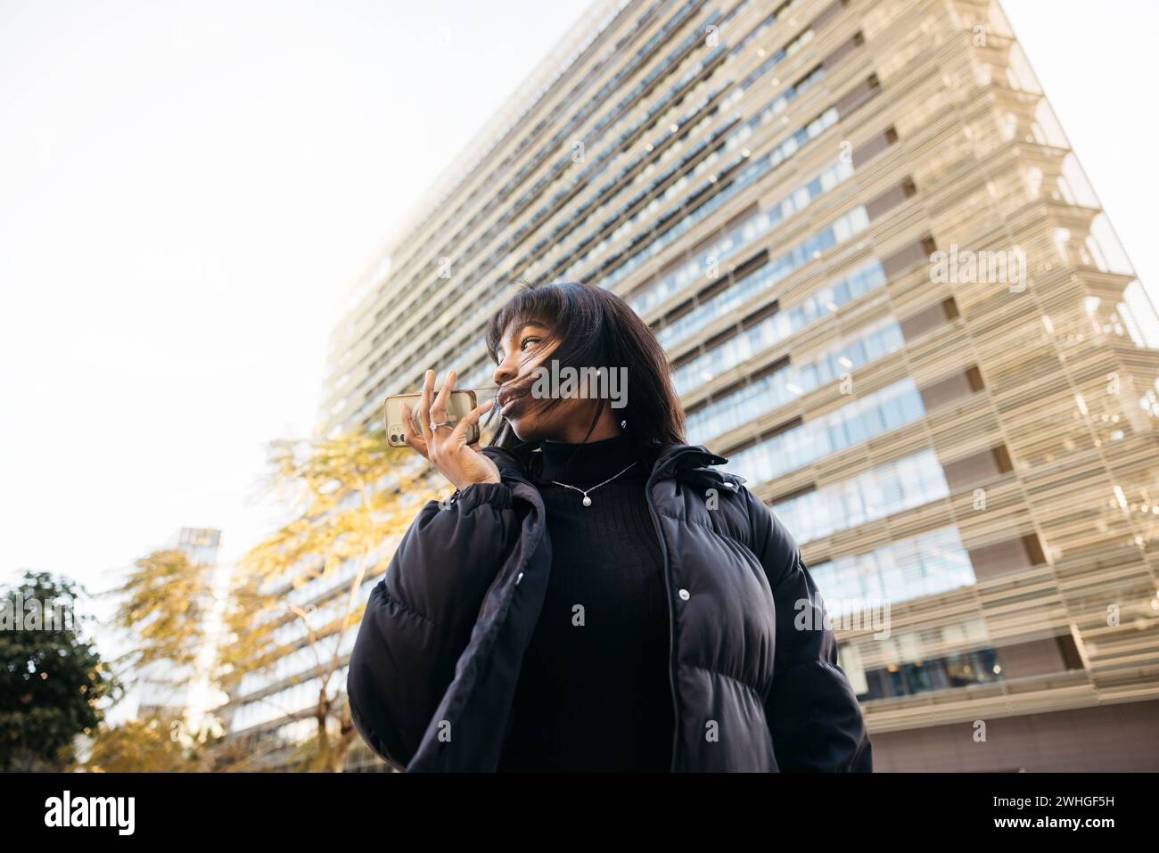 Junge schöne schwarze Frau in schwarzer Kleidung, die eine Sprachnachricht auf einem Telefon sendet, während sie auf der Straße läuft. Stockfoto