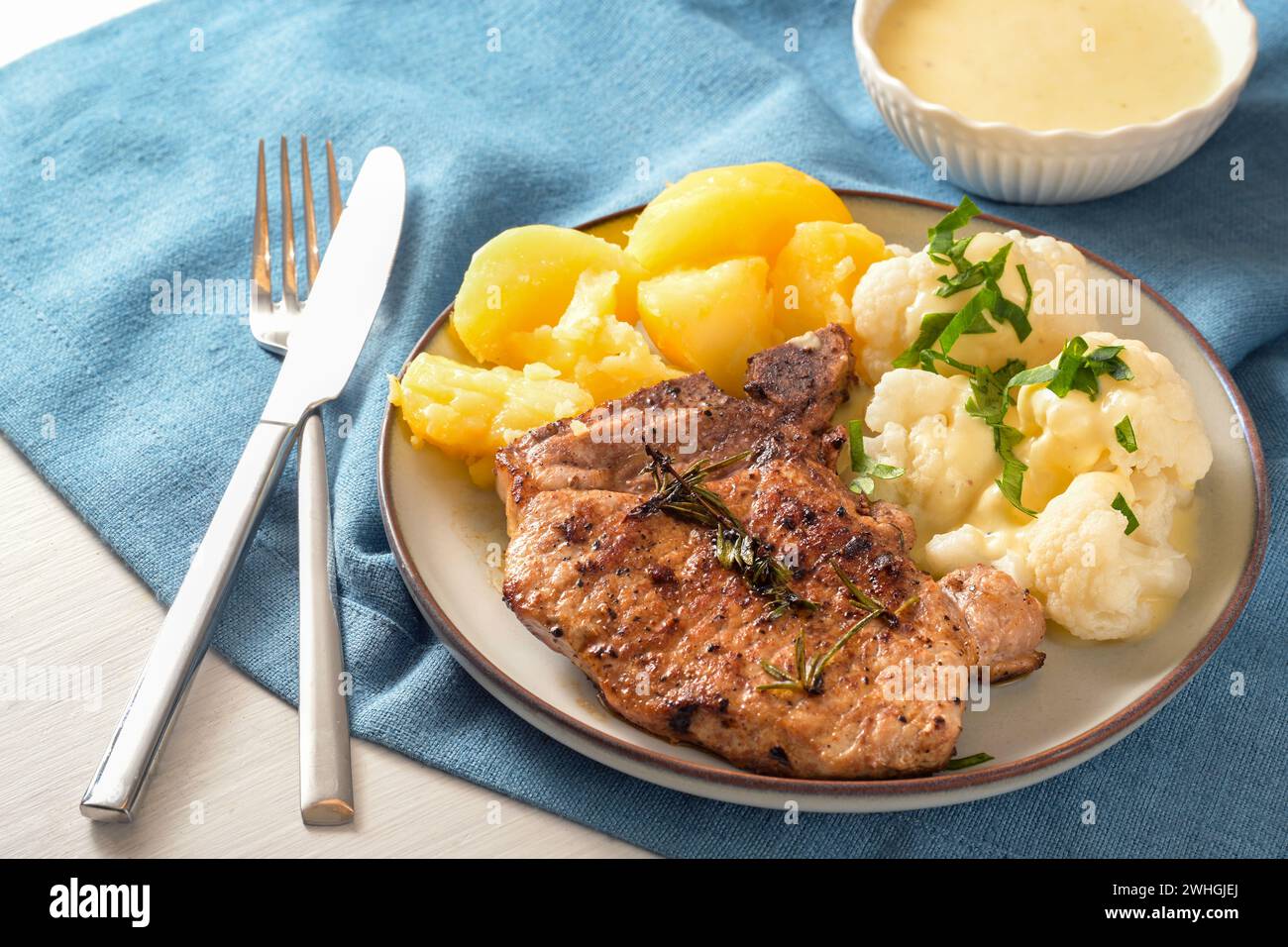 Gericht mit Schweinekotelett, Blumenkohl, Kartoffeln und Hollandaise-Sauce serviert mit Kräutergarnitur auf einem Teller und einer blauen Serviette, ausgewählt Stockfoto