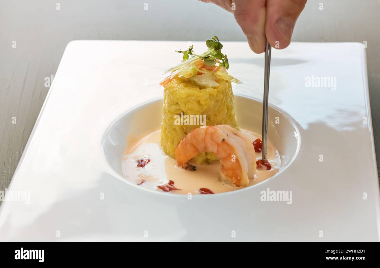 Hand mit einer Pinzette garnieren Sie ein Gourmet-Gericht aus Safranrisotto mit Kräuterbelag, einer großen Garnelen- und Krustentiersauce in einem Mod Stockfoto