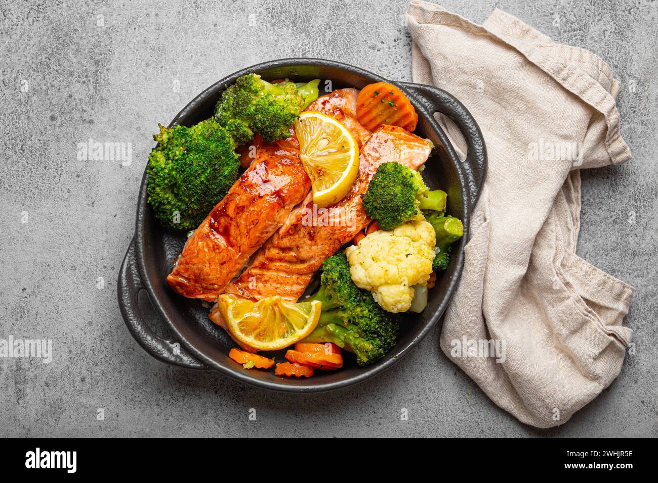 Gesunde gebackene Fisch Lachssteaks, Brokkoli, Blumenkohl, Karotte in schwarzem gusseisernen Auflaufschüssel auf grauem rustikalem Steinhintergrund Stockfoto