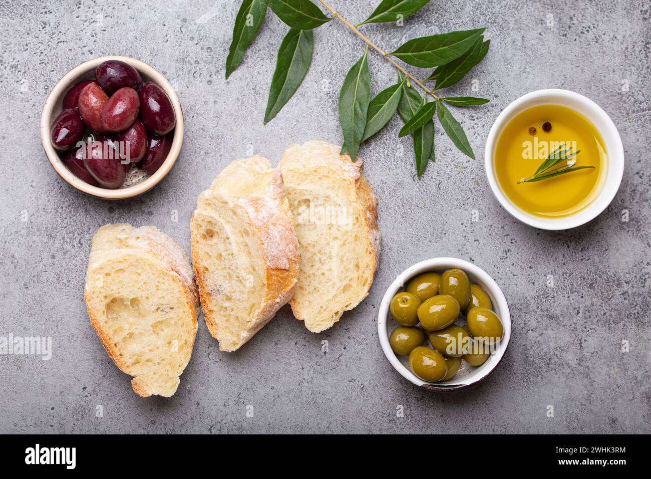 Frische Ciabatta-Scheiben, grüne und braune Oliven, Olivenöl mit Rosmarin, Olivenzweige auf grauem Betonstein rustikale bac Stockfoto