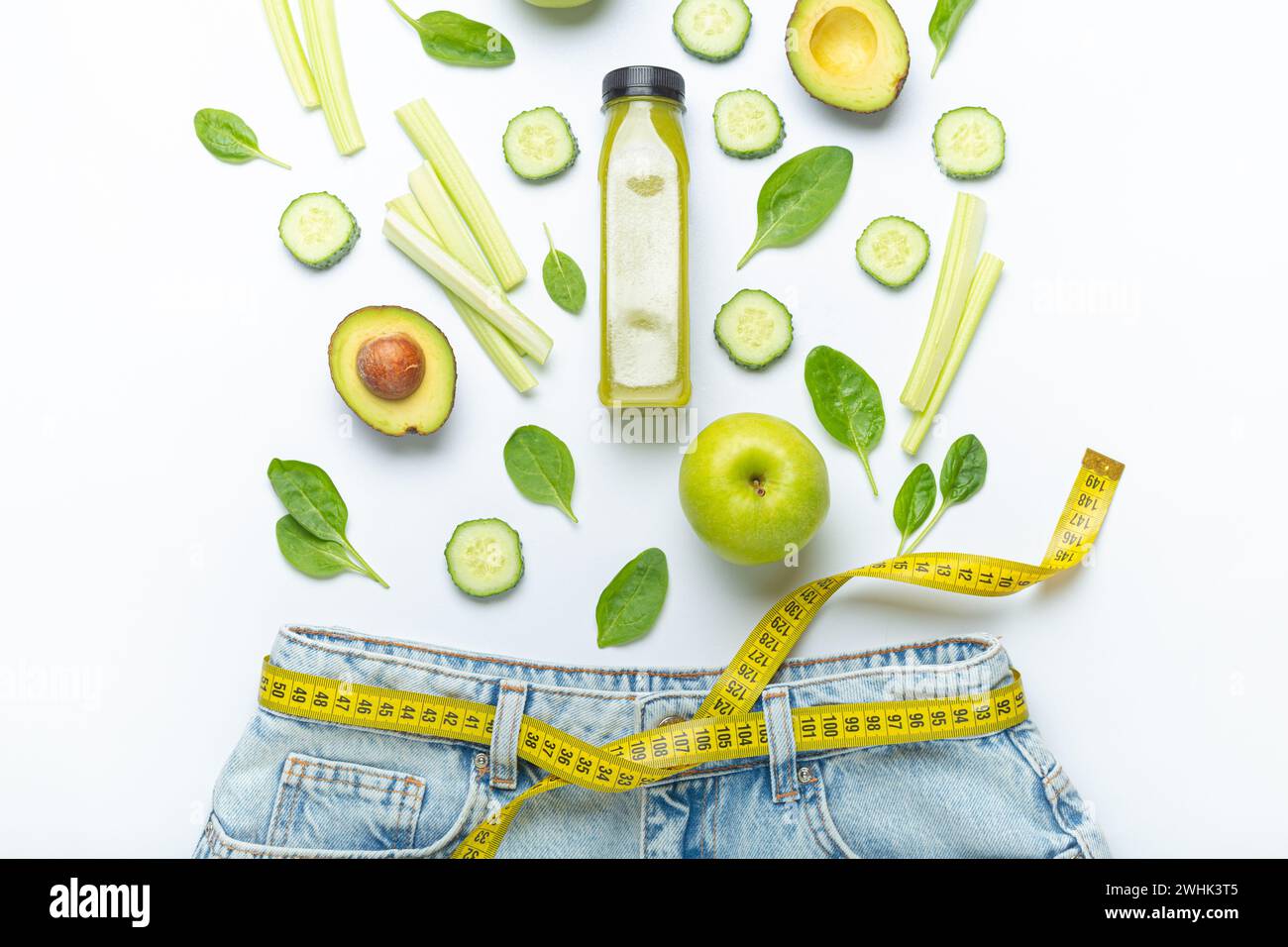 Grünes Obst, Gemüse, Smoothie, der in Jeans fällt, und gelbes Maßband statt Gürtel auf weißem Hintergrund. Konzept von W Stockfoto