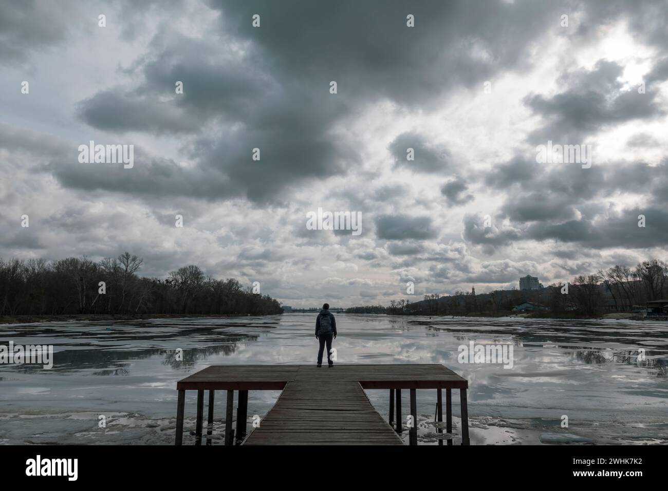 Einsame Frau steht auf dem Holzsteg und sieht Wolkenreflexionen im kalten Wasser. Dramatisches Foto über harte Zeiten, schwierige Lebensentscheidungen und d Stockfoto