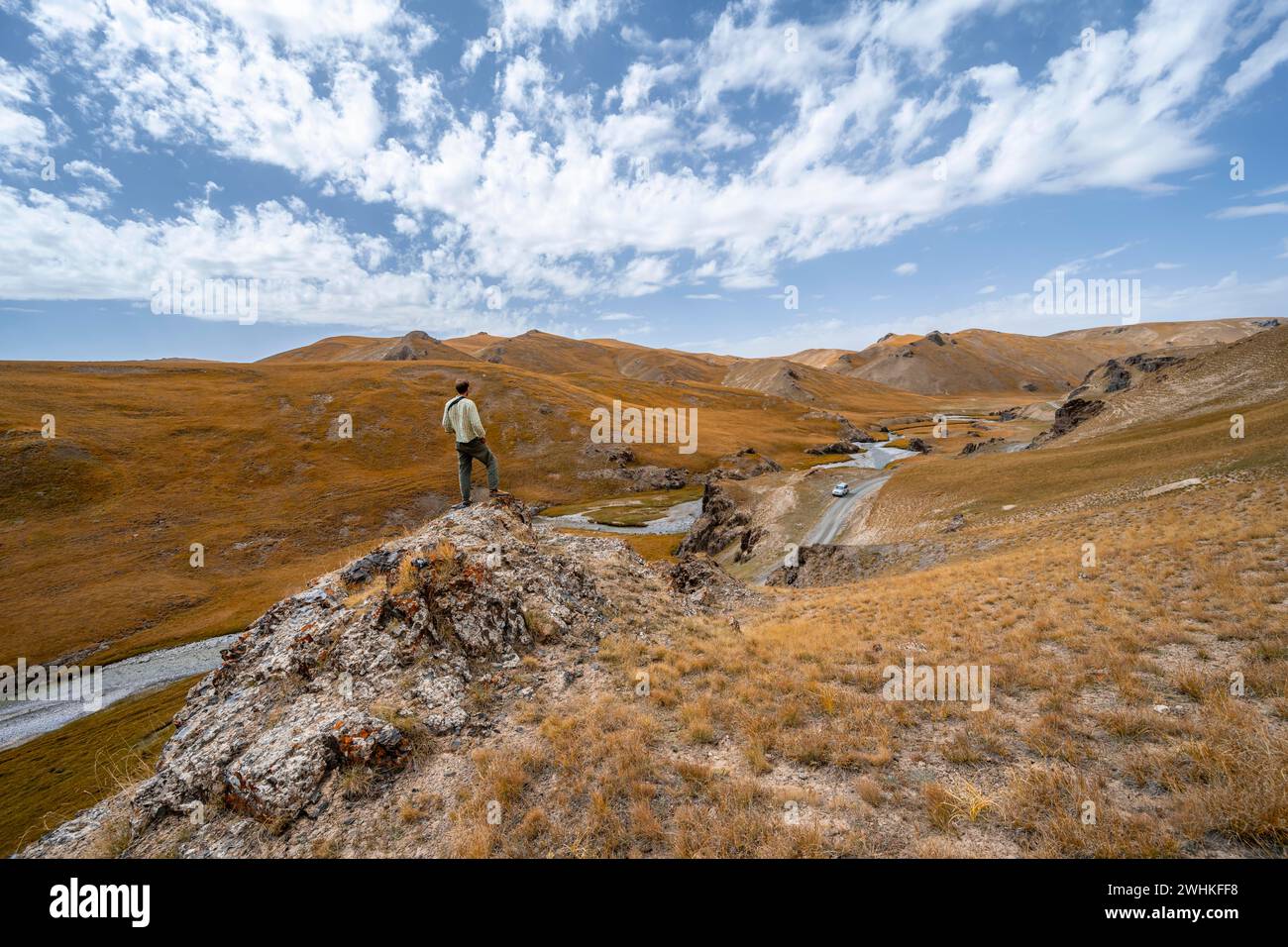 Wanderer auf einem Felsen, Auto auf der Straße durch Bergtal mit Hügeln mit gelbem Gras, Provinz Naryn, Kirgisistan Stockfoto