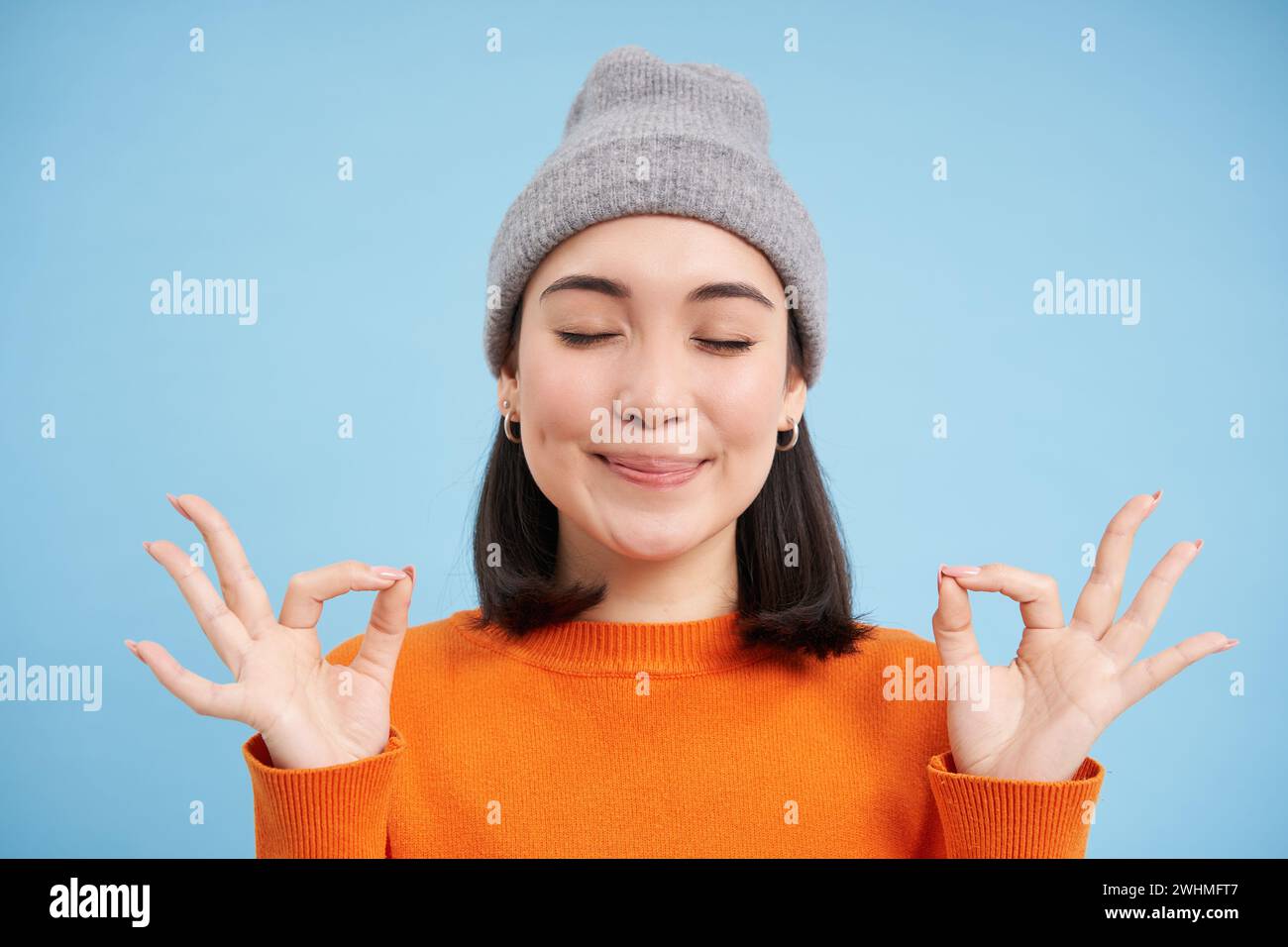 Klingt Geist im gesunden Körper. Lächelndes ruhiges und entspanntes asiatisches Mädchen in Mütze, zeigt Zen, Entspannungsgeste, Meditation, Stehen Stockfoto