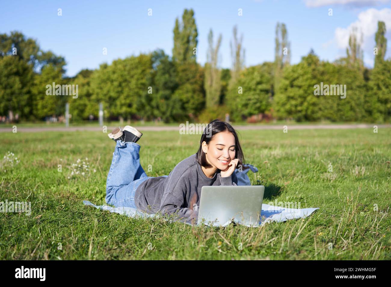 Porträt eines jungen, glücklichen asiatischen Mädchens, das im Park auf einer Decke liegt, Videos ansieht und auf ihrem Laptop im Internet surft Stockfoto