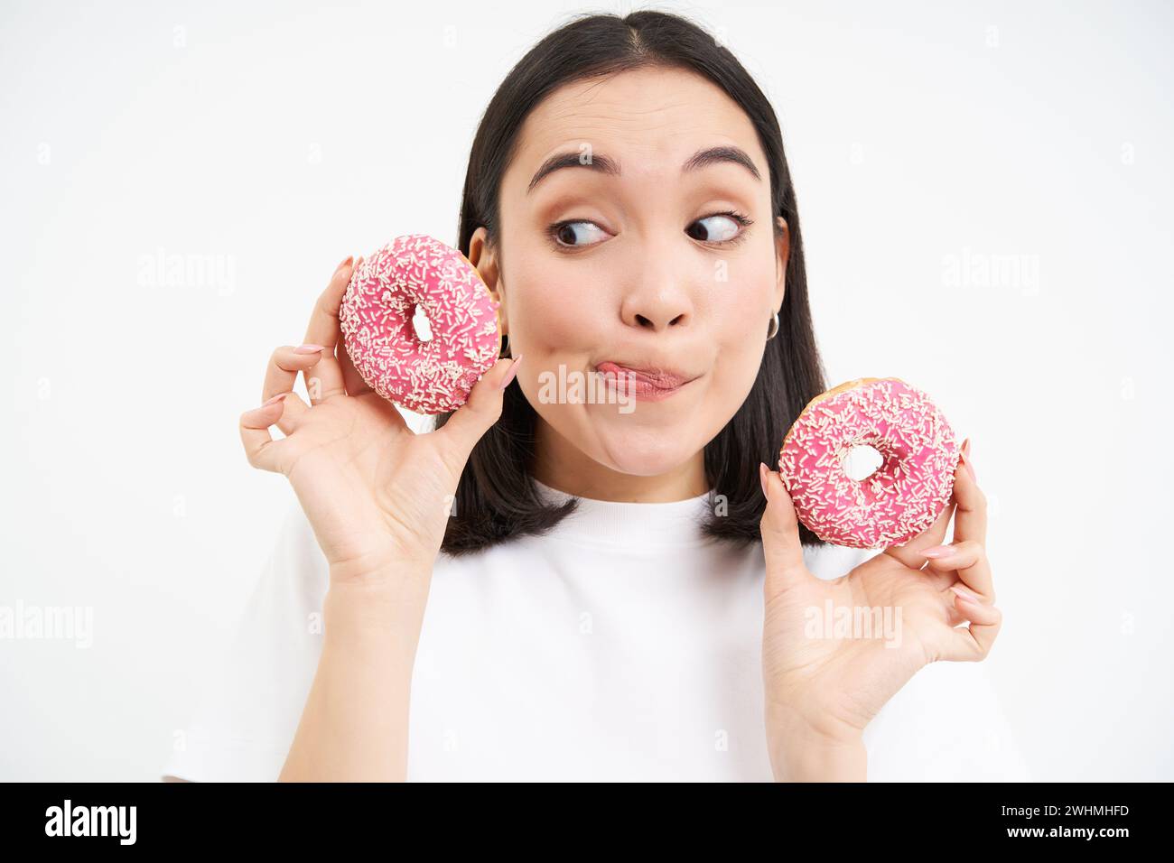 Porträt einer lustigen asiatischen Frau, die versucht war, leckere glasierte Dougnut zu essen, zwei Donuts in der Hand zu halten und aufgeregt lächelnd, isoliert auf weißem B Stockfoto