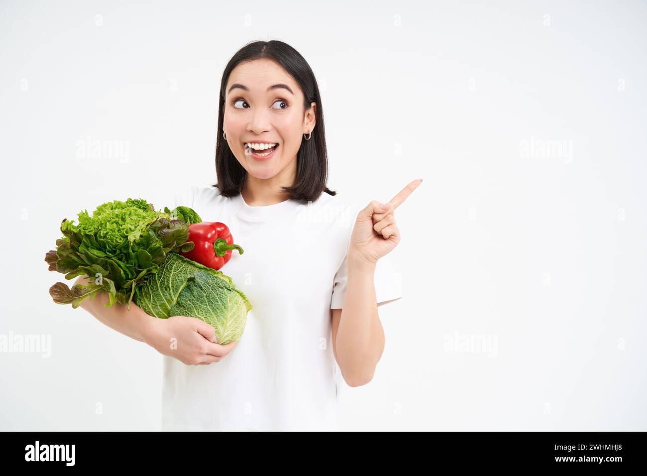 Enthusiastische koreanerin, die ein paar Gemüse hält, mit dem Finger direkt auf das Banner zeigt, Werbewerbung zeigt, weißer Rücken Stockfoto