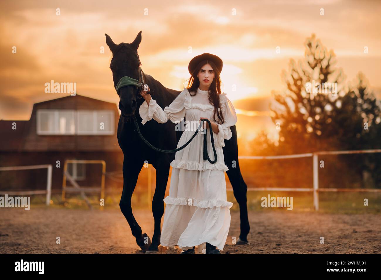 Frau in weißem Kleid und schwarzem Hut mit ihrem Pferd bei Sonnenuntergang draußen auf der Ranch. Stockfoto