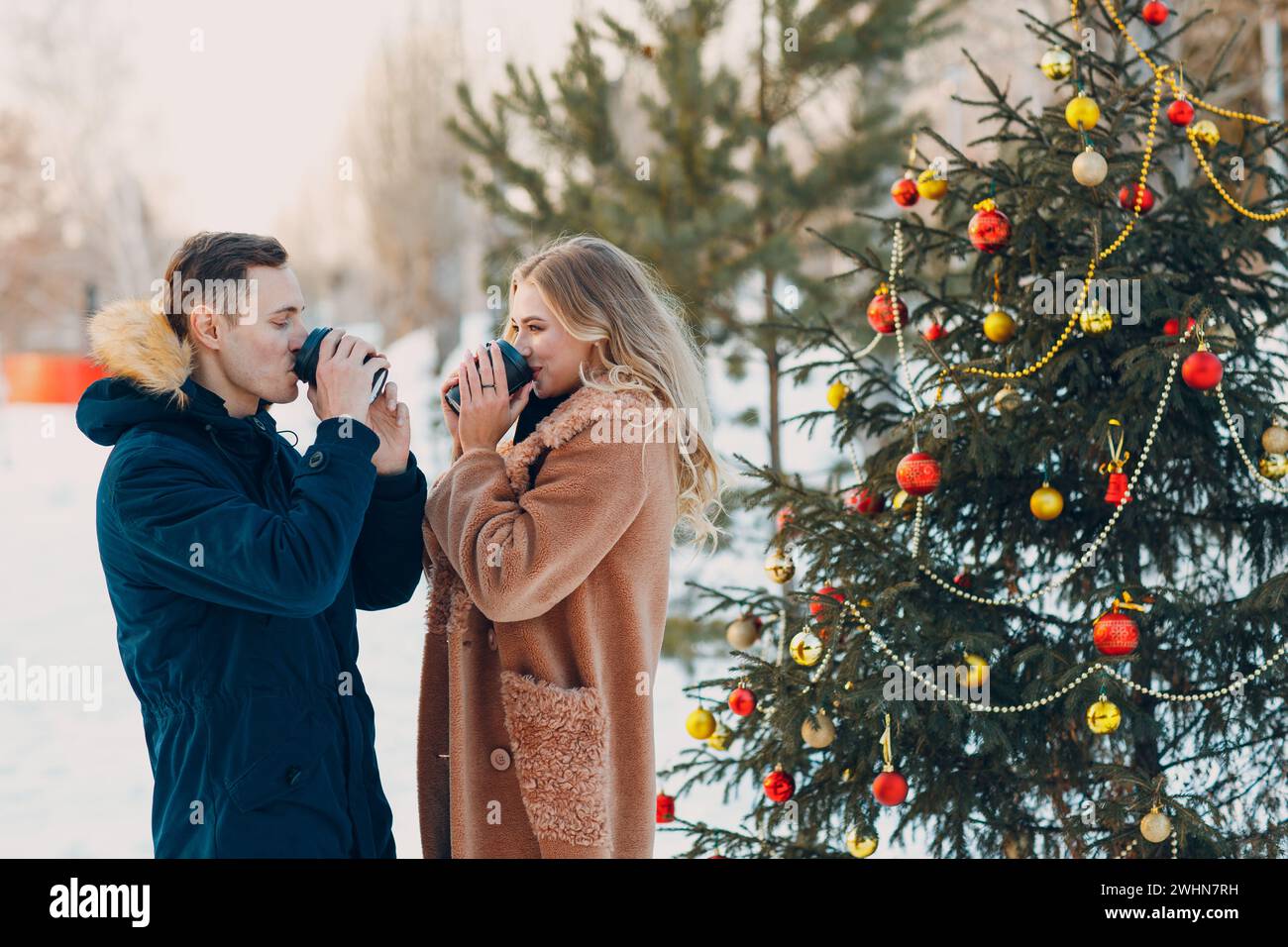 Verliebte Paare trinken Kaffee neben dem geschmückten Weihnachtsbaum im Winterwald Stockfoto