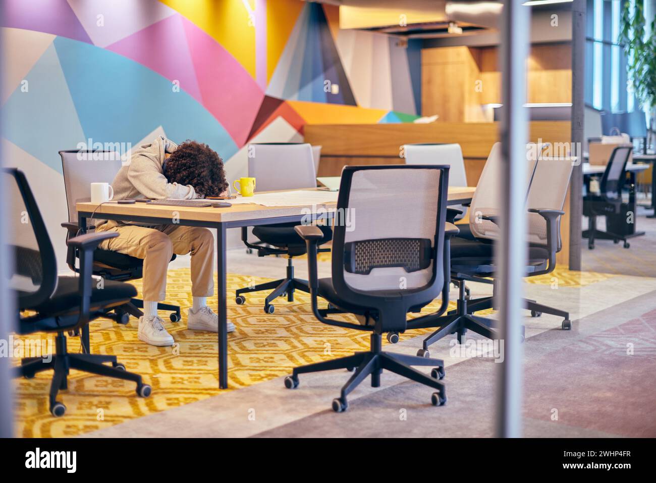 In einem modernen, geräumigen Startup-Büro liegt ein erschöpfter Afroamerikaner auf einem Schreibtisch und spiegelt die Herausforderungen des Zeitgenossen wider Stockfoto