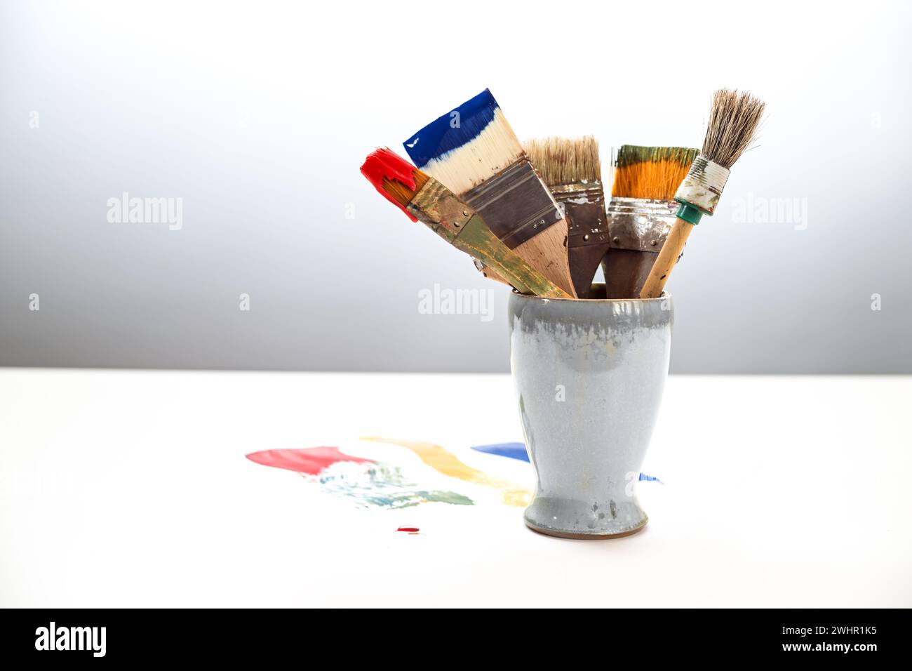 Mehrere benutzte Pinsel mit verschiedenen Farben in einem Keramikbecher und Farbflecken auf dem weißen Tisch vor grauem Hintergrund, Stockfoto