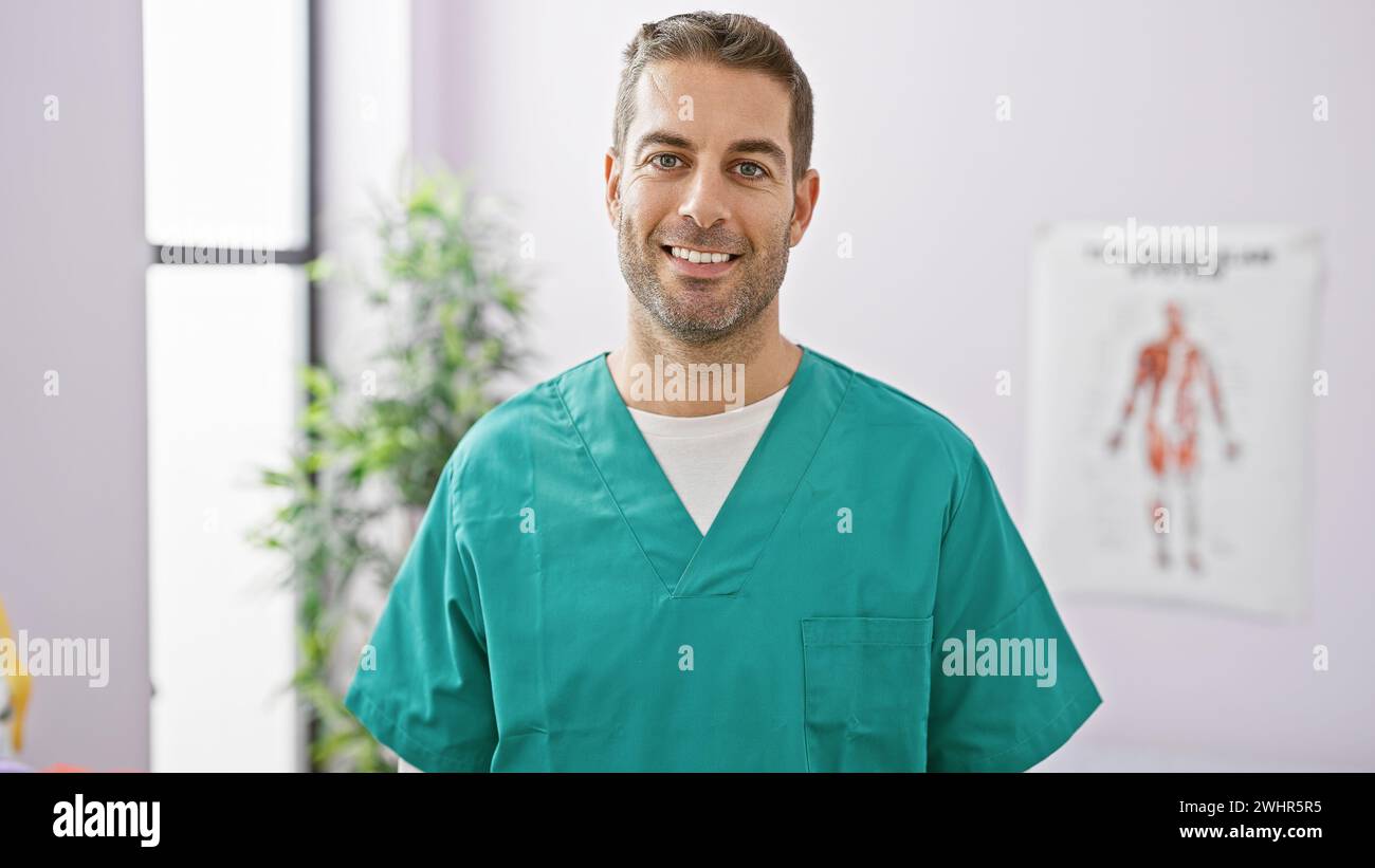 Ein lächelnder hispanischer Mann in grüner Schale, der selbstbewusst in einem sauberen Krankenhauszimmer mit einem Poster des Bewegungsapparates im Hintergrund steht. Stockfoto