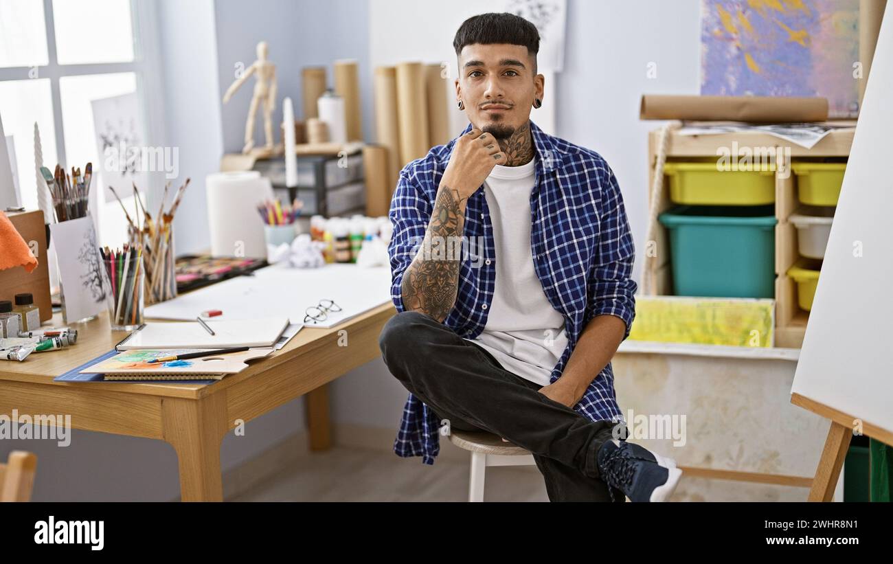 Tätowierter junger lateinamerikanischer Mann, ein vertiefter Künstler, der mit ernstem Gesicht in einem Kunststudio an seiner Staffelei sitzt, mit Pinsel in der Hand Stockfoto
