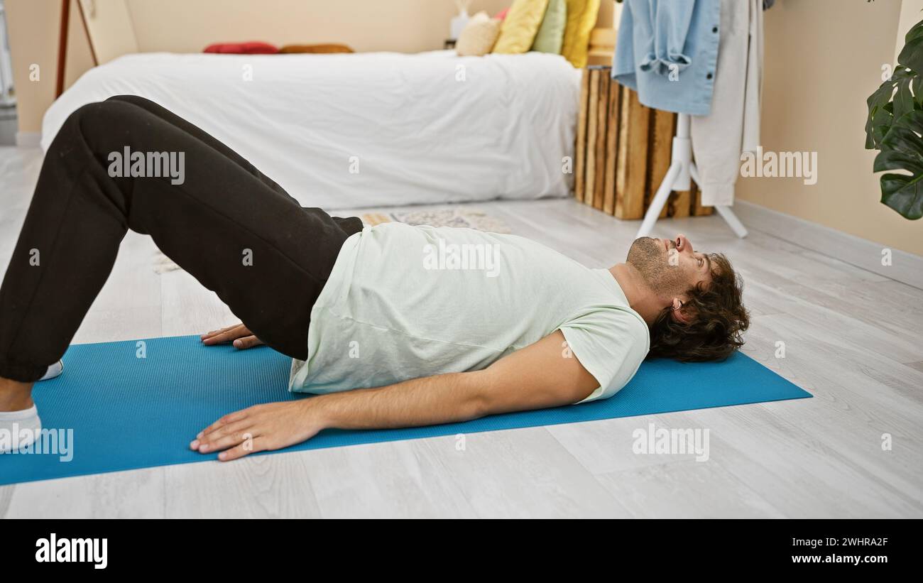 Ein junger bärtiger Mann entspannt sich auf einer Yogamatte in einem hellen Schlafzimmer mit moderner Einrichtung und Zimmerpflanzen, die Wellness und Ruhe verkörpern. Stockfoto