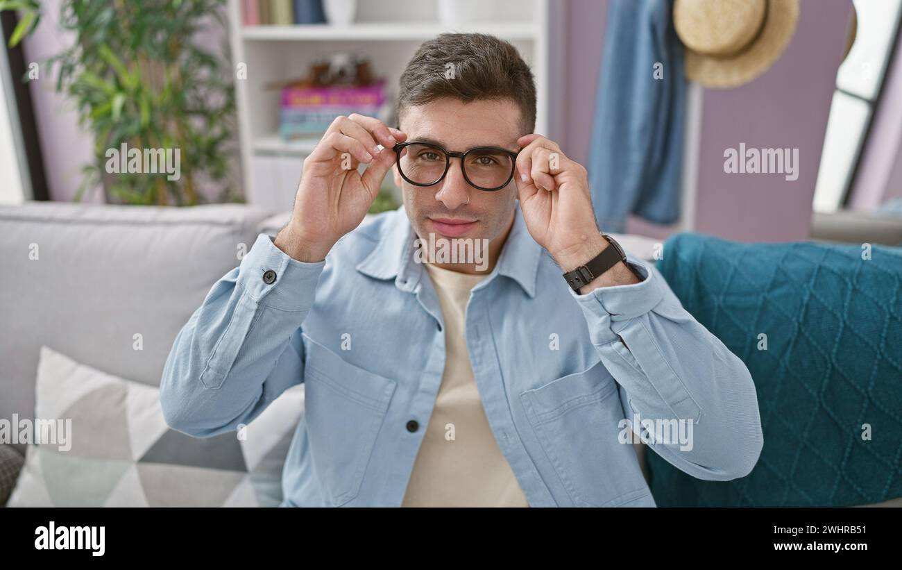 Fröhlicher, gutaussehender junger hispanischer Mann mit einem selbstbewussten Lächeln, der sich bequem auf dem Sofa im Wohnzimmer zu Hause entspannt und dabei die Gemütlichkeit im Innenraum genießt Stockfoto