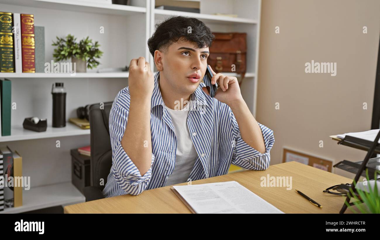 Ein junger Mann in einem gestreiften Hemd telefoniert an einem organisierten Schreibtisch mit Bücherregalen im Hintergrund. Stockfoto