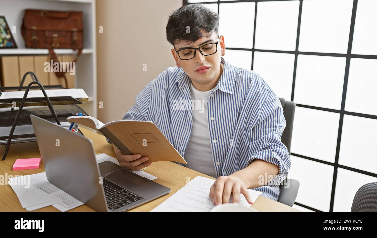 Ein junger Mann in Brille liest ein Notizbuch an einem überfüllten Schreibtisch mit einem Laptop und Dokumenten. Stockfoto