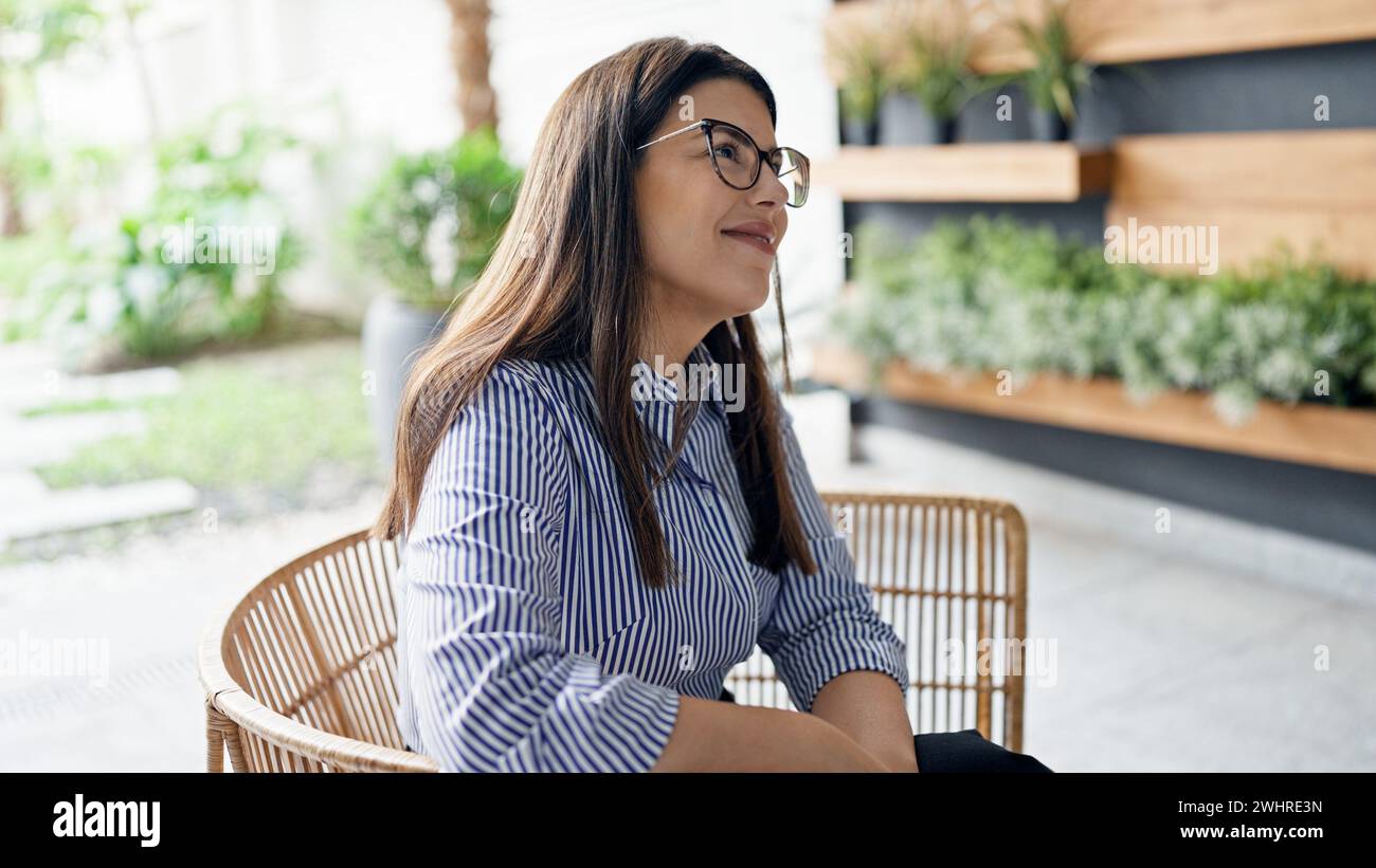 Junge, schöne hispanische Frau, die selbstbewusst lächelnd auf einem Stuhl im Bürogarten sitzt Stockfoto