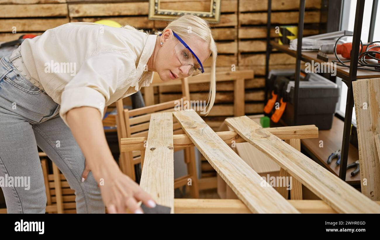 Die junge blonde Zimmerfrau trägt eine Sicherheitsbrille, die Holzplanken bei der Zimmerei schleift Stockfoto