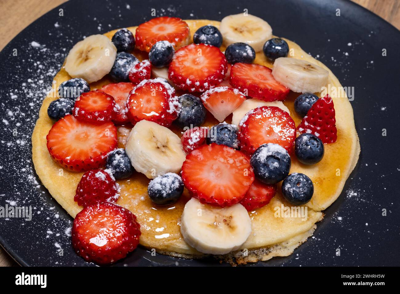 Schließen Sie das Frühstück mit Pfannkuchen mit Obst, Erdbeeren, Bananen und Heidelbeeren mit Ahornsirup und Puderzucker, glutenfrei Stockfoto