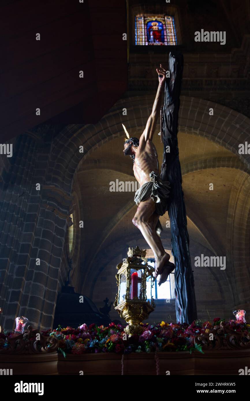 Eine Skulptur der Kreuzigung, die von natürlichem Licht beleuchtet wird und sich im dunklen Inneren einer gotischen Kirche erhebt, umgeben von bunten Blumen in t Stockfoto