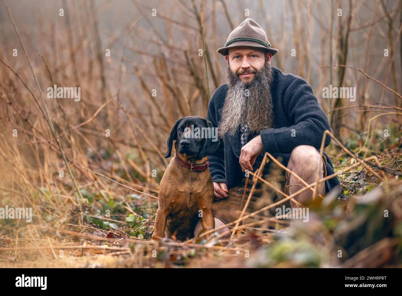 Ein bayerischer Mann in traditioneller Tracht, der mit seinem bayerischen Berghund in der Nähe interagiert Stockfoto