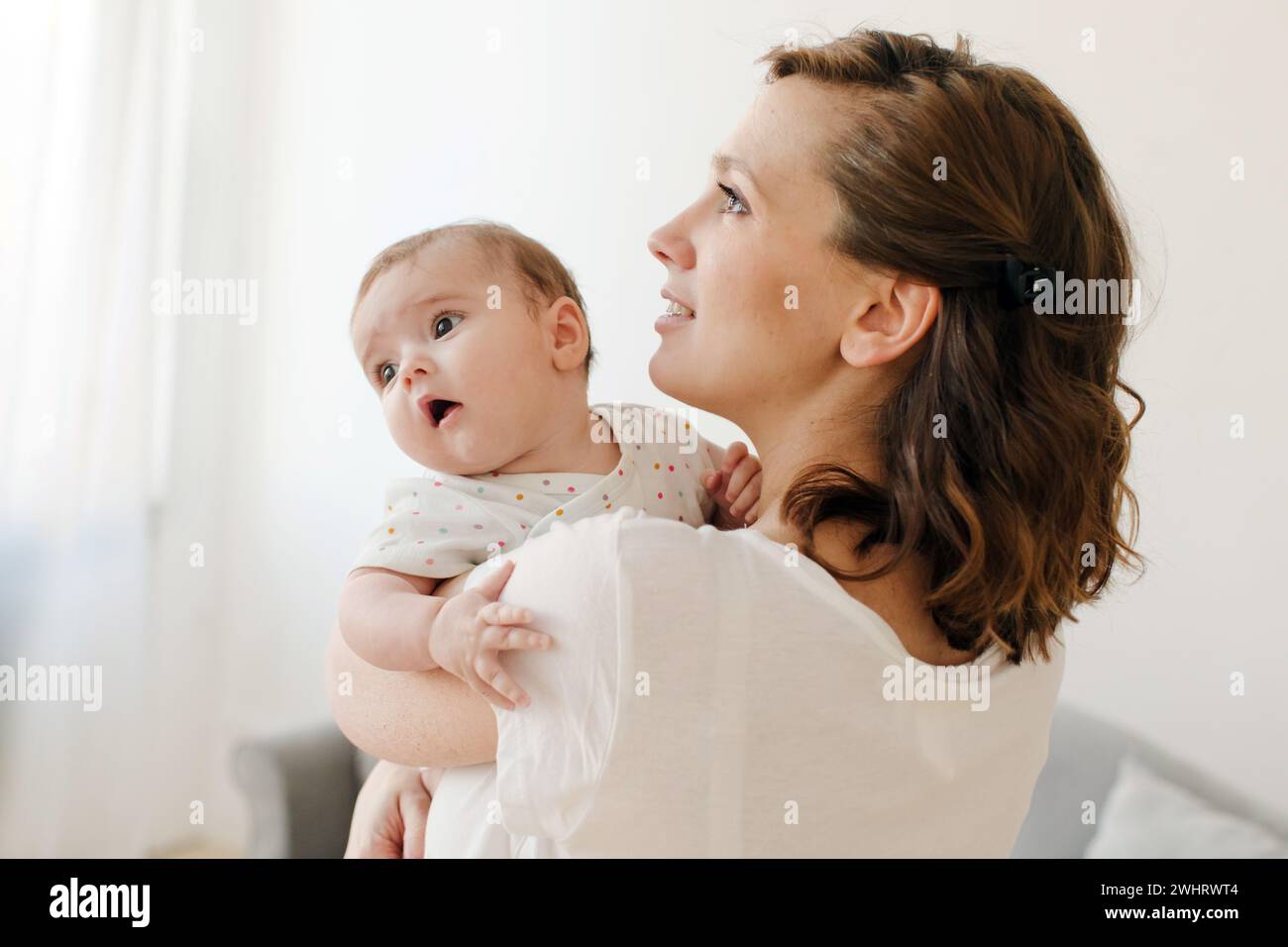 Seitenansicht einer fröhlichen jungen brünetten Frau in lässiger Kleidung, die ein wertvolles Kleinkind im sonnendurchfluteten Wohnzimmer umschließt und nach oben blickt Stockfoto