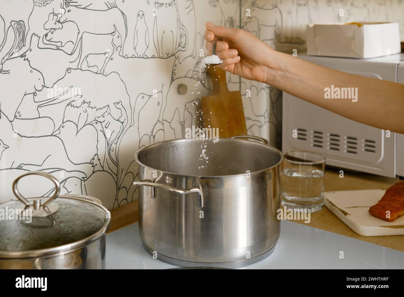 Nicht erkennbare Frau benutzt Löffel, um Salz in einen Topf mit kochendem Wasser auf dem Elektroherd zu geben Stockfoto
