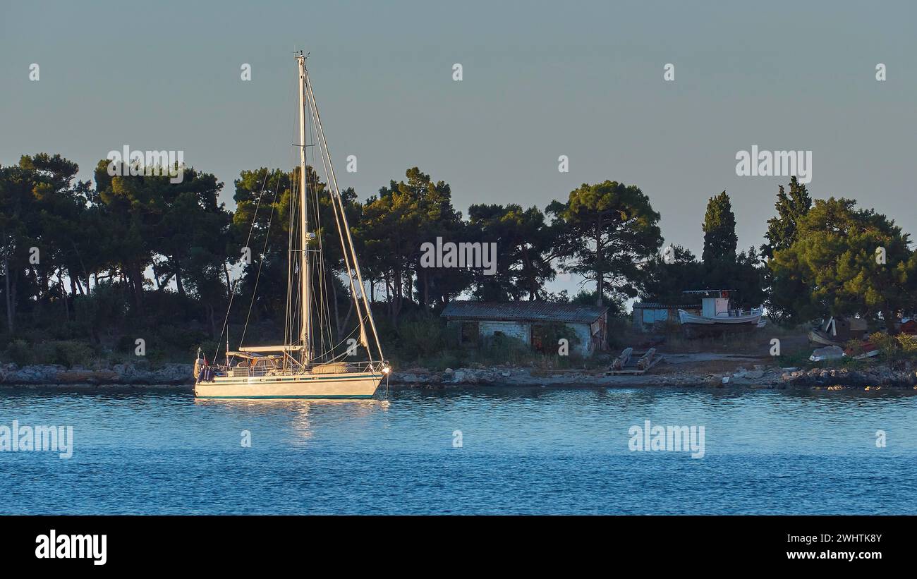 Ein Segelboot schwimmt bei Sonnenaufgang in der Nähe der Küste mit Bäumen, Gythio, Mani, Peloponnes, Griechenland Stockfoto