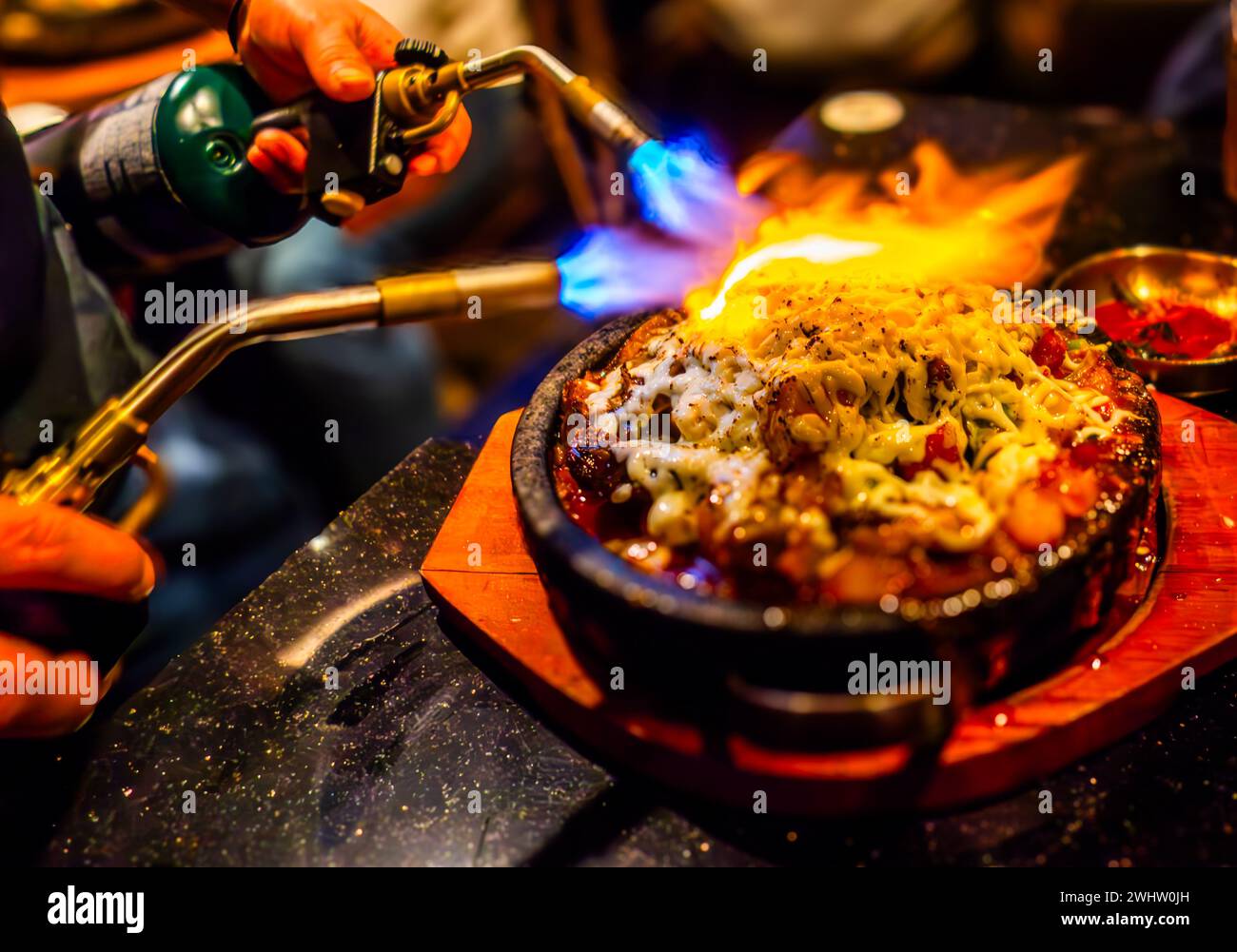 Eine Person, die ein Gericht auf einem Herd mit sichtbaren Flammen in einer Schüssel kocht Stockfoto