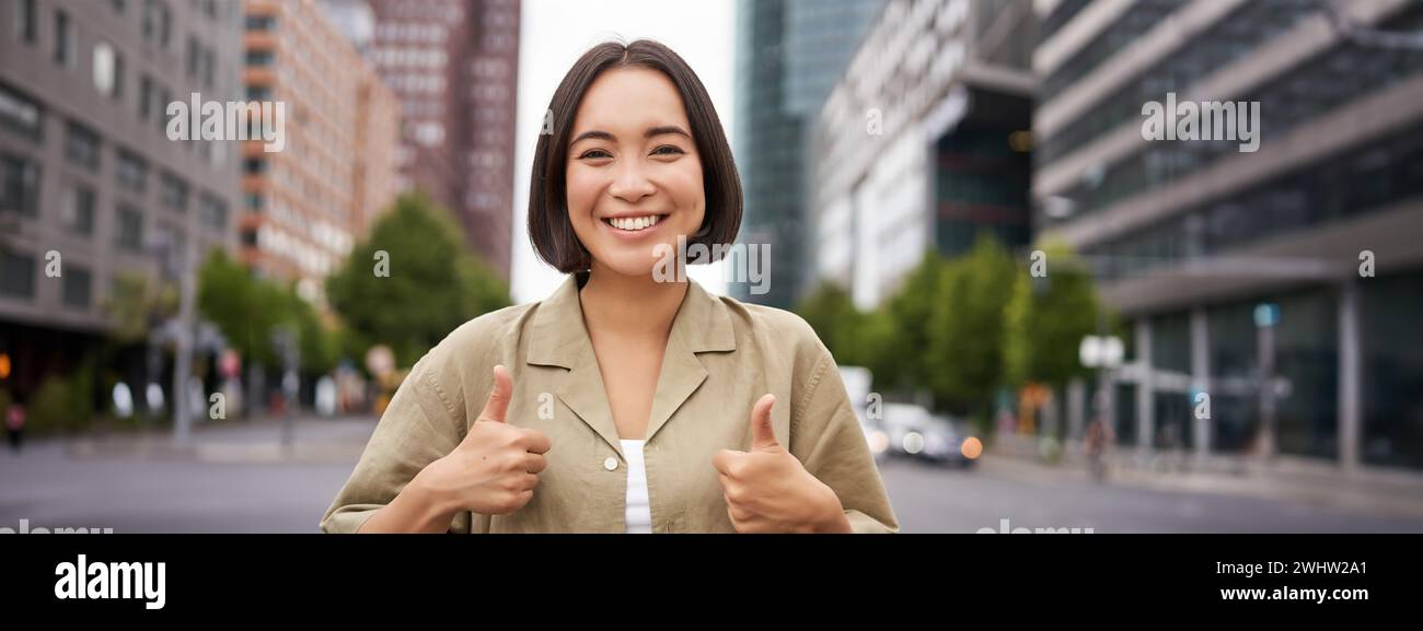 Enthusiastische asiatische Frau, zeigt Daumen nach oben in Zustimmung, schaut optimistisch, sagt ja, stimmt zu und stimmt zu, steht auf der Straße Stockfoto