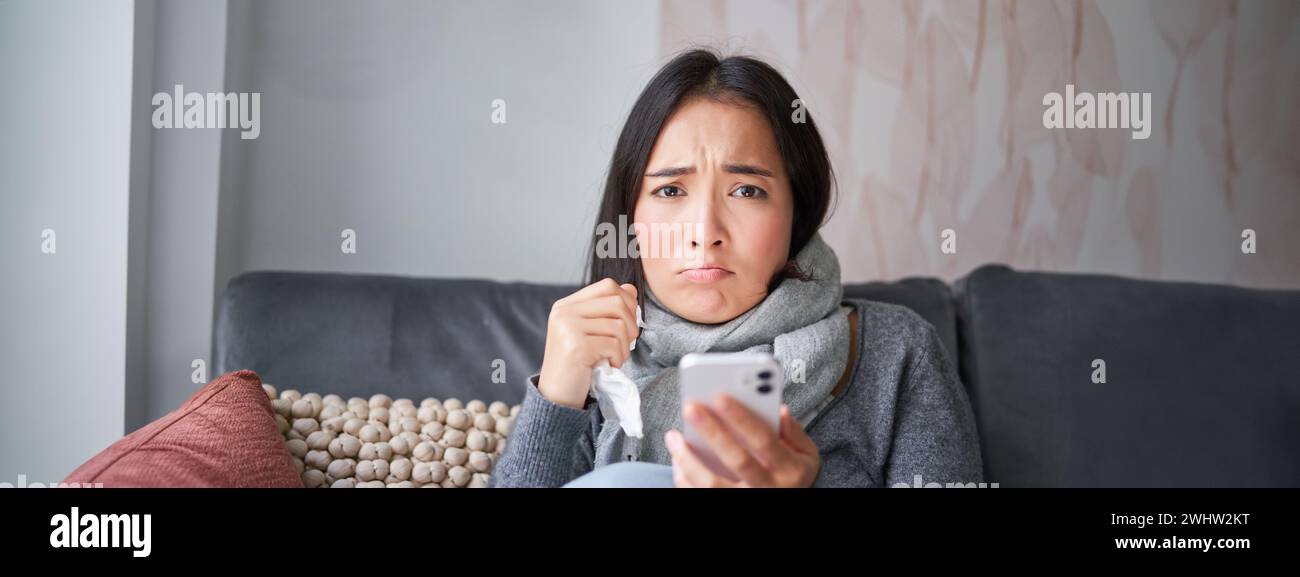 Asiatisches Mädchen mit Grippe, sitzt zu Hause, weint und sieht verärgert aus, hält das Smartphone, fühlt sich unwohl und düster, trägt warme Kleidung Stockfoto