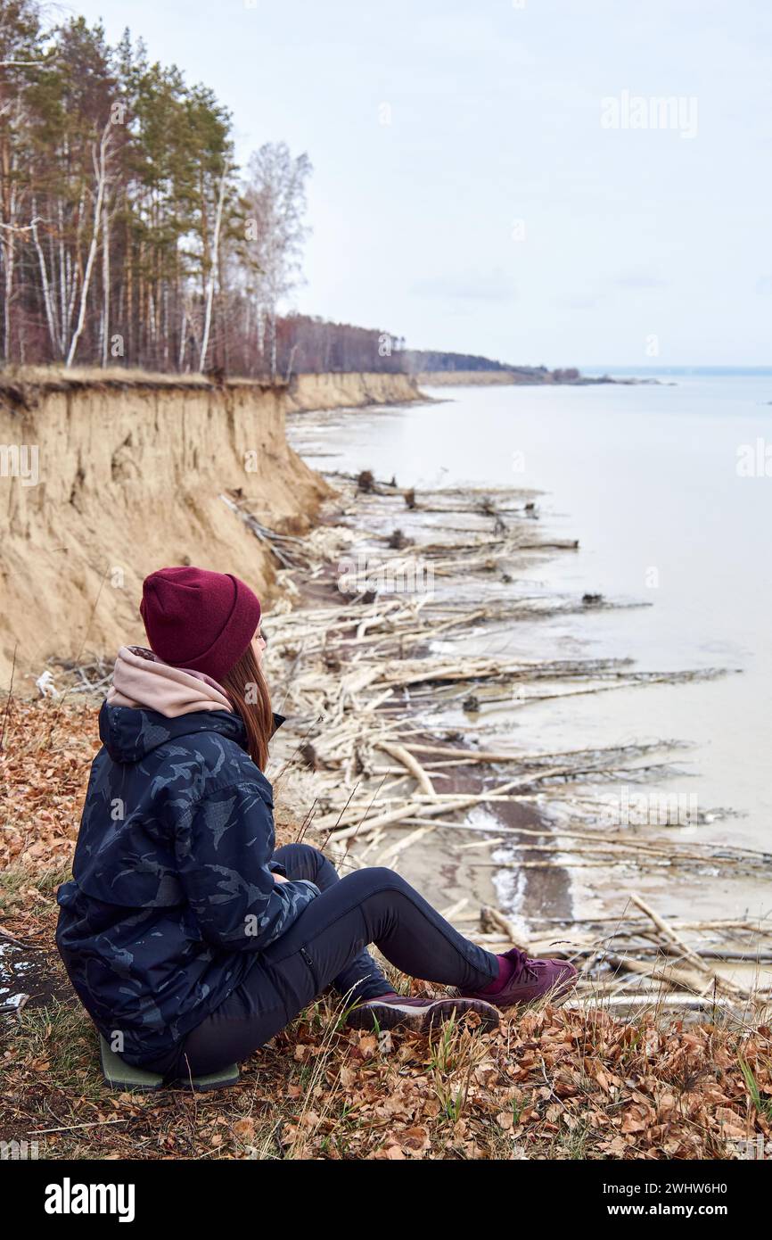 Frau in warmen Kleidern, sitzt auf einer Klippe über der Küste, bewundert die Natur, genießt die Einsamkeit. Aktiver Lifestyle-Moment. Das Konzept, bei jedem Wetter zu reisen Stockfoto