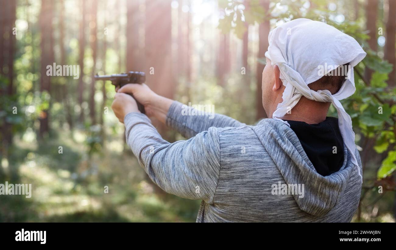 Ein Mann schießt eine Pistole im Wald zwischen den Bäumen, die von Sonnenlicht beleuchtet werden. Rückansicht, unscharfer Hintergrund. Stockfoto