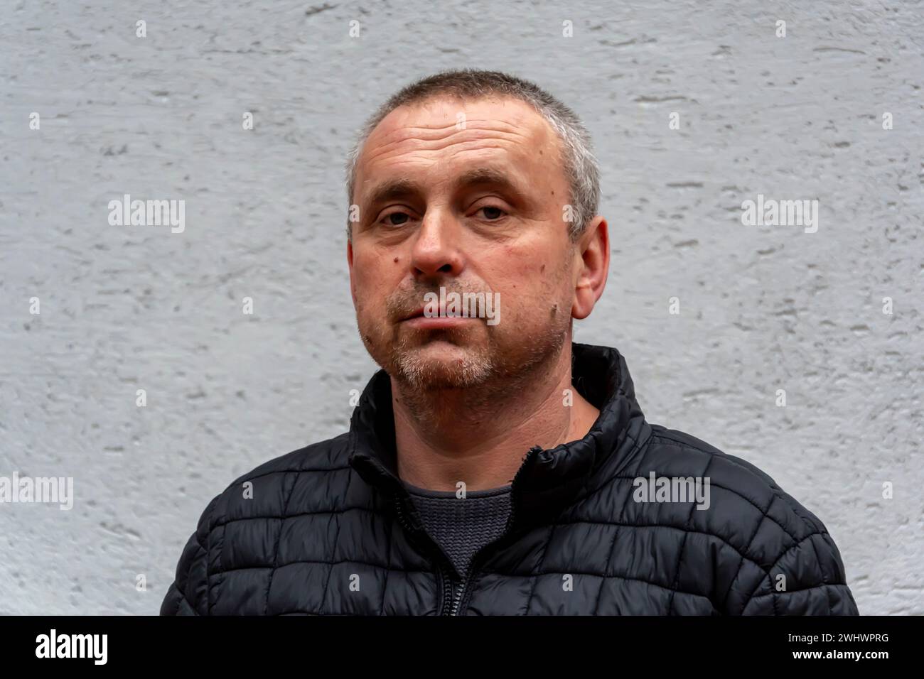 Ein grauhaariger älterer Mann, 45-50 Jahre alt, an einer hellen Wand, der eine schwarze Jacke trägt, schaut in die Kamera. Stockfoto