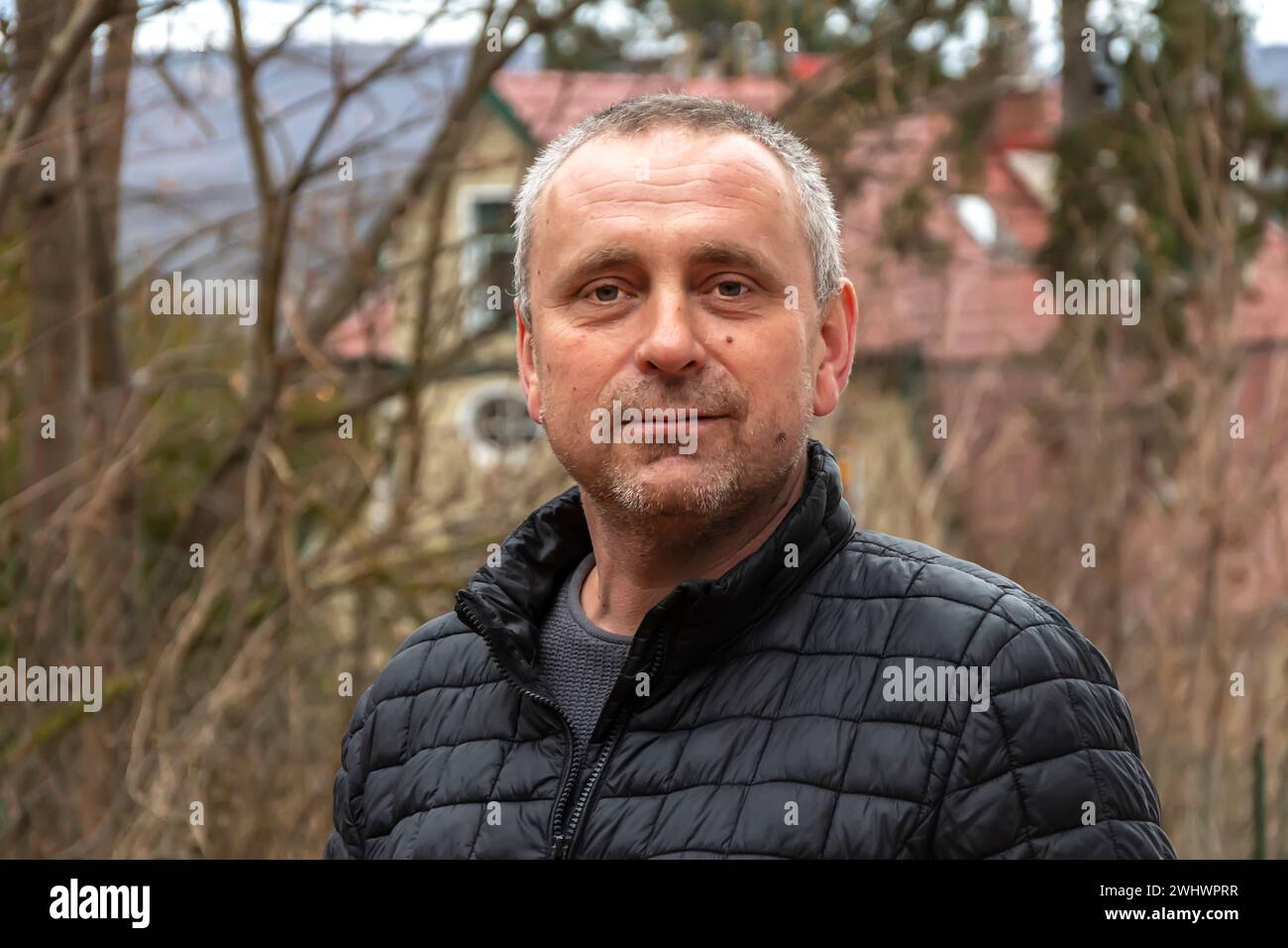 Grauhaariger Mann 45-50 Jahre alt in einer schwarzen Jacke auf dem Hintergrund eines Landhauses und eines Waldes. Stockfoto