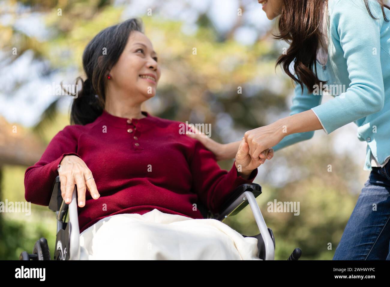 Familienbeziehung Asiatische Seniorin im Rollstuhl mit glücklicher Tochter, die eine Pflegeperson um die Hand hält, während sie Zeit mit dem Kind verbringt Stockfoto