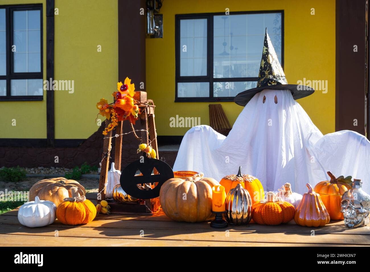 Kind im Bettlaken mit Schlitzen wie Geist in Hexenhut und Halloween Dekor auf der Veranda des Hauses draußen im Hof von Pum Stockfoto