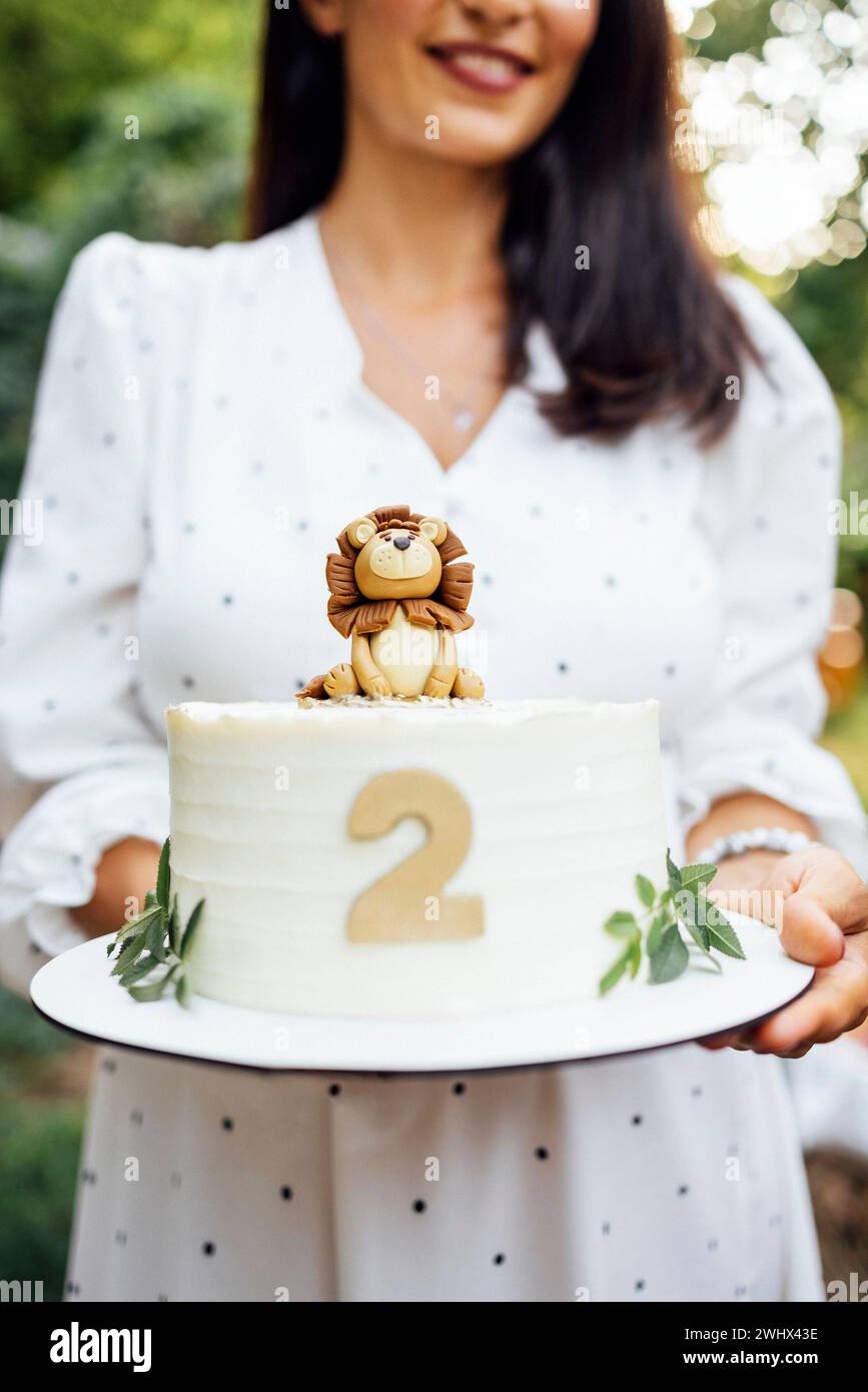 Nahaufnahme von weiblichen Händen halten einen Geburtstagskuchen. Köstlicher süßer Kuchen mit niedlichem Löwenjungen, Nummer zwei und Blättern. Stockfoto