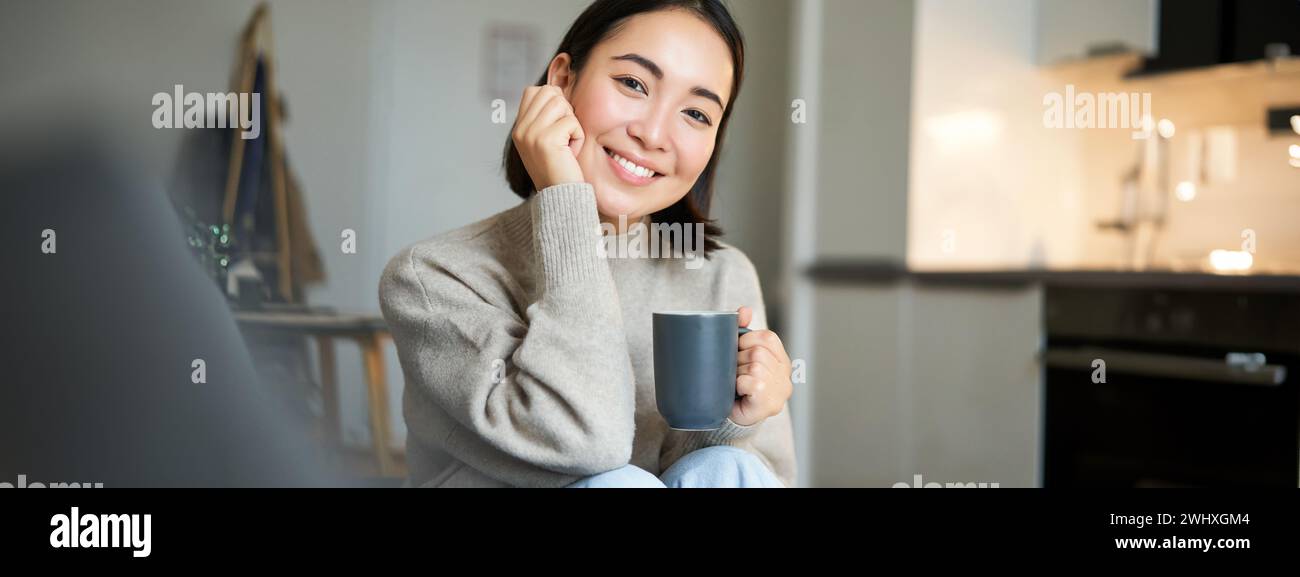 Lächelnde asiatische Frau sitzt zu Hause mit einer Tasse Kaffee, entspannt und fühlt sich warm, blickt aus dem Fenster, ruht auf dem Sofa Stockfoto